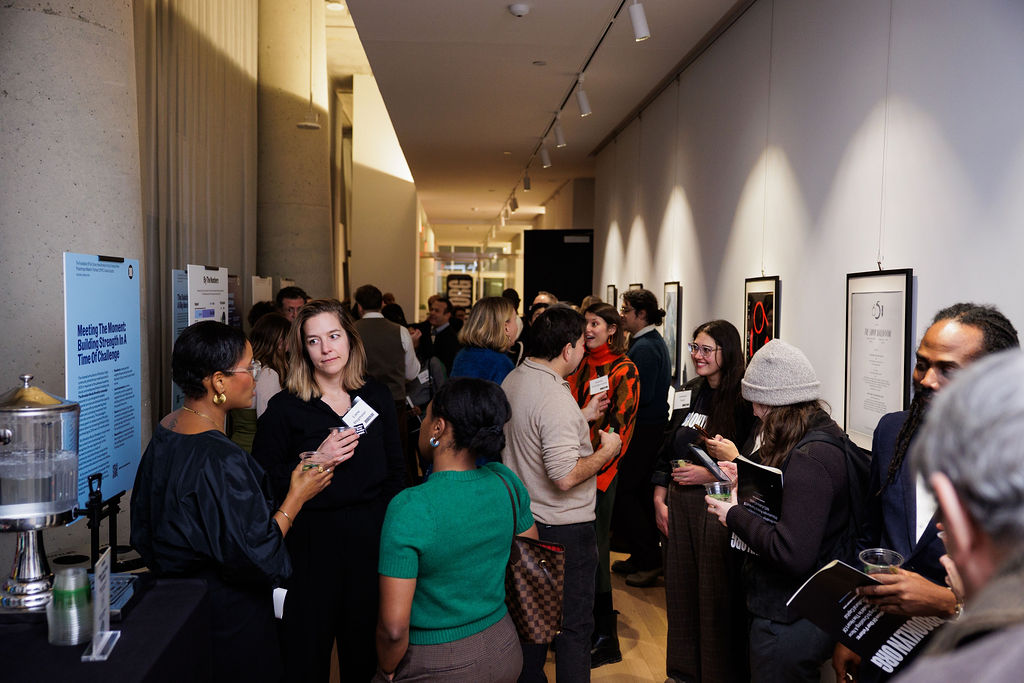 A group of people converse and network in a hallway at an indoor event, with informational posters and framed artwork displayed on the walls.