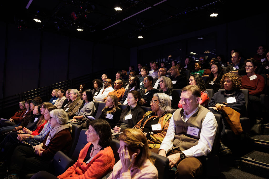 A diverse group of people sits in tiered rows of seats, attentively watching a presentation or performance in a dimly lit auditorium.