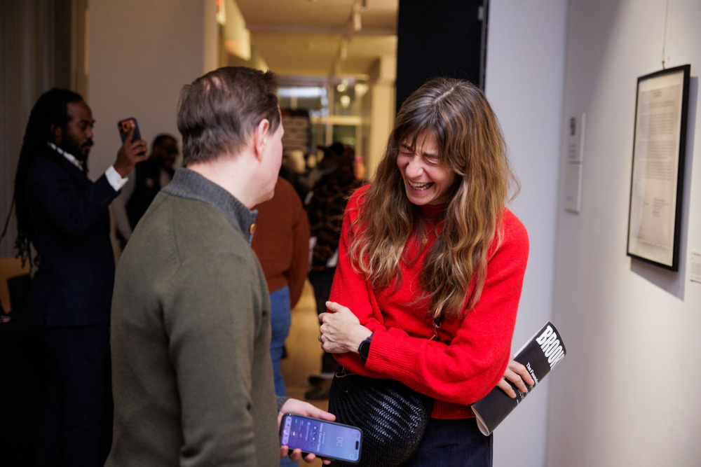 A woman in a red sweater laughs while talking to a man holding a phone at an indoor event; other people are visible in the background.