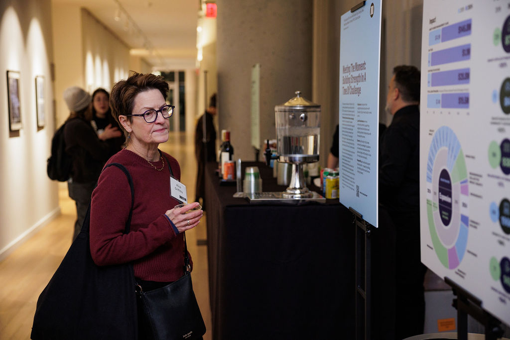 A person with short hair and glasses looks at an informational display in a hallway with other people and refreshments in the background.
