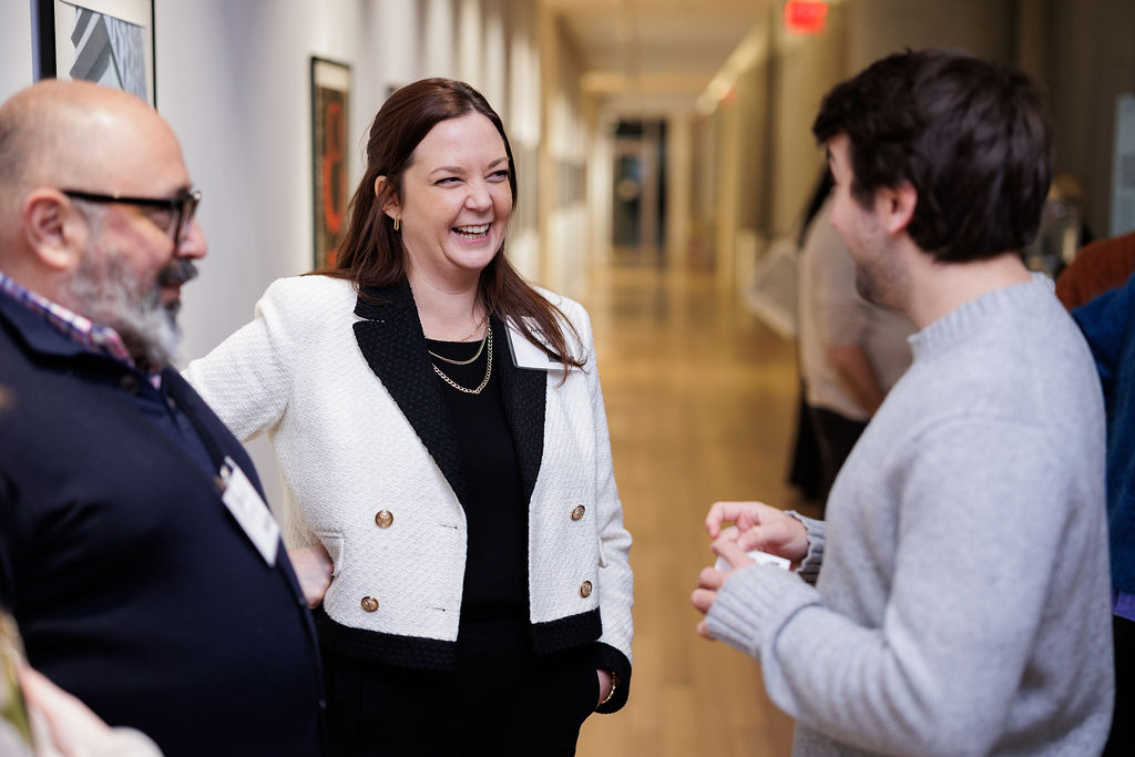 Three people stand in a hallway having a conversation. The woman in the center is smiling and talking, while the two men listen. The background is a blurred corridor with wall art and lights.