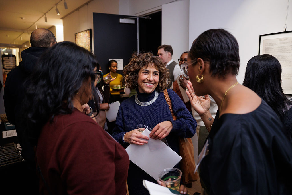 A group of people stand and converse at an indoor event; one woman in the center is smiling and holding a drink while others listen and engage.