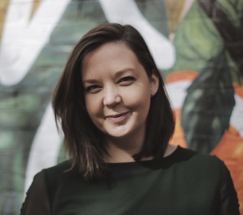 A woman with shoulder-length brown hair smiles slightly while standing in front of a colorful, abstract mural.