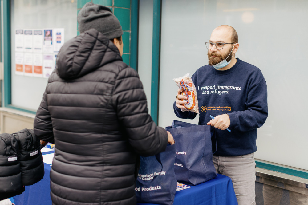 A man wearing a sweatshirt that says "I support immigrants and refugees" gives a packaged loaf of bread to someone at an outdoor distribution table.