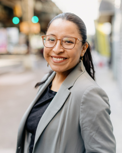 A woman with glasses and long dark hair, wearing a light gray blazer and black top, smiles while standing outdoors on a city street.