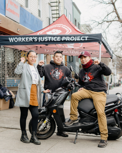 Three people pose with raised fists in front of a tent labeled "Worker's Justice Project"; one sits on a motor scooter while the other two stand on either side.