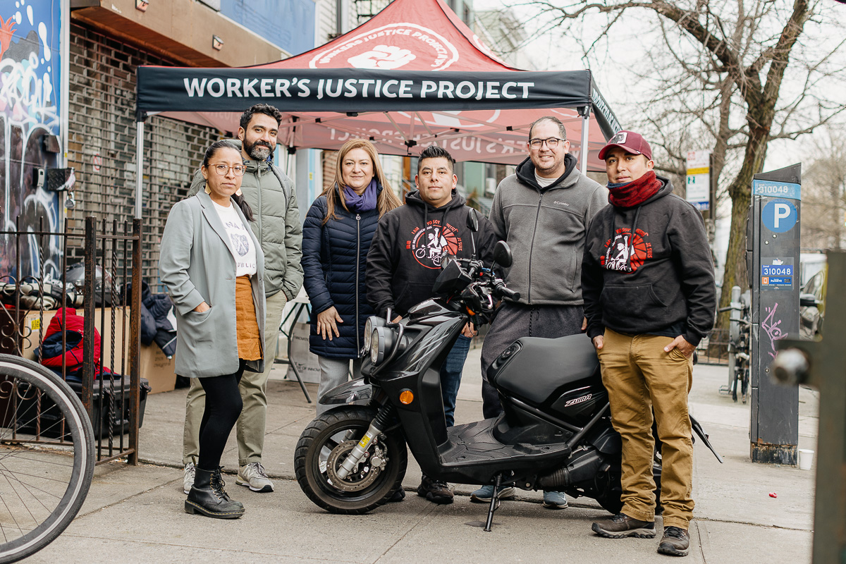 Six people stand in front of a "Worker's Justice Project" tent on a city sidewalk, posing beside a parked scooter.