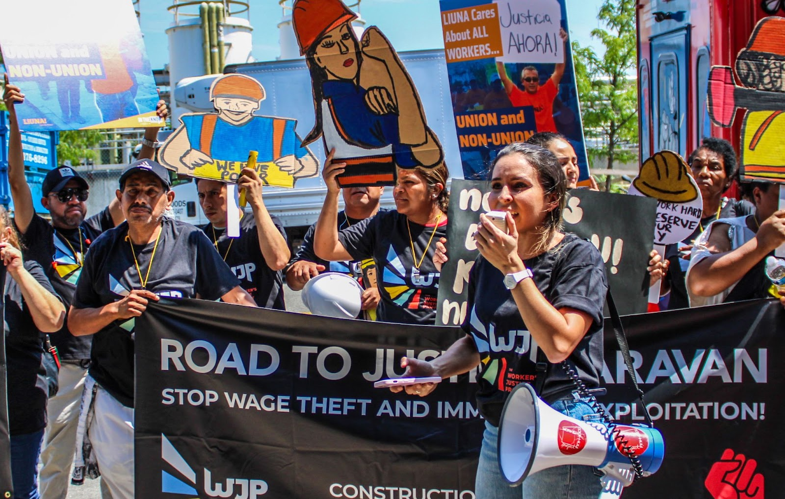 A group of protesters march holding signs and a banner reading "Road to Justice Caravan: Stop Wage Theft and Immigrant Exploitation!" One person speaks into a megaphone.