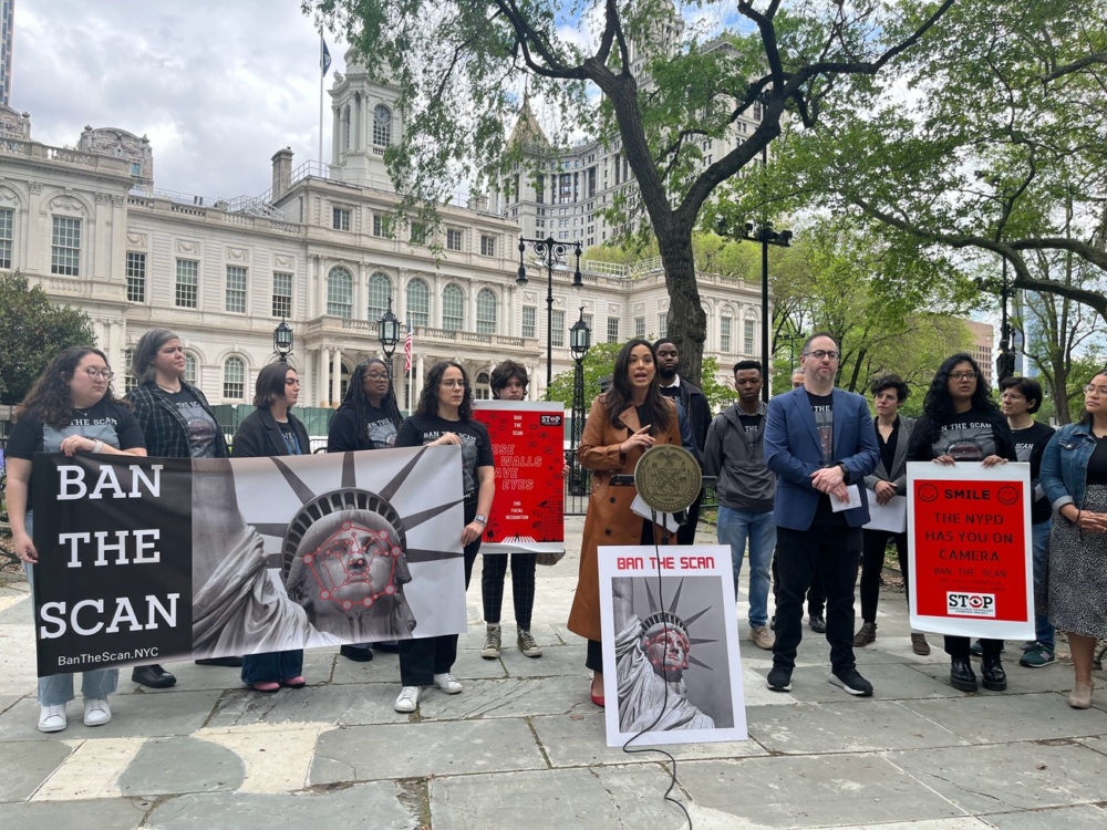 A group of people stand outside a government building holding signs protesting facial recognition surveillance, including one that says "BAN THE SCAN.