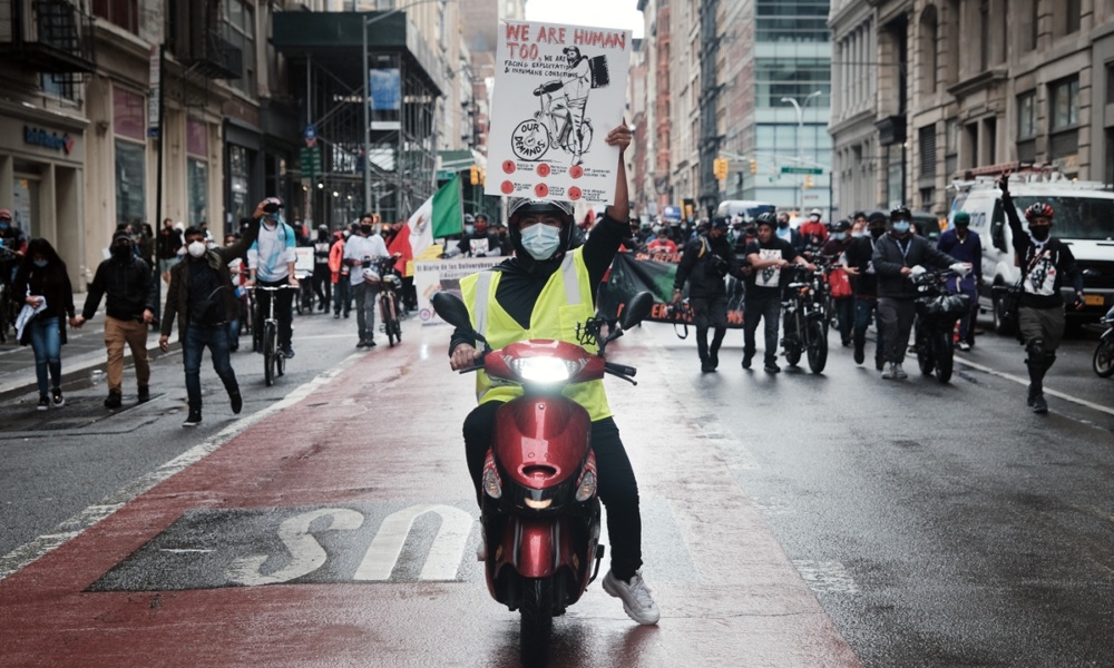 A person wearing a yellow vest and mask rides a scooter, holding a protest sign, leading a group of people marching on a city street.