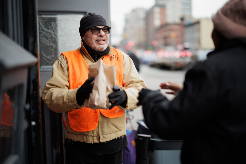 A person wearing an orange safety vest hands a paper bag to another person outdoors in an urban setting.