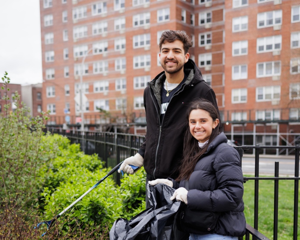 Two people wearing gloves pick up litter with a trash bag and grabber tool near bushes in front of an apartment building.