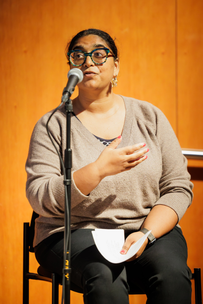A person wearing glasses and a beige sweater speaks into a microphone while seated, holding a piece of paper against an orange-brown background.