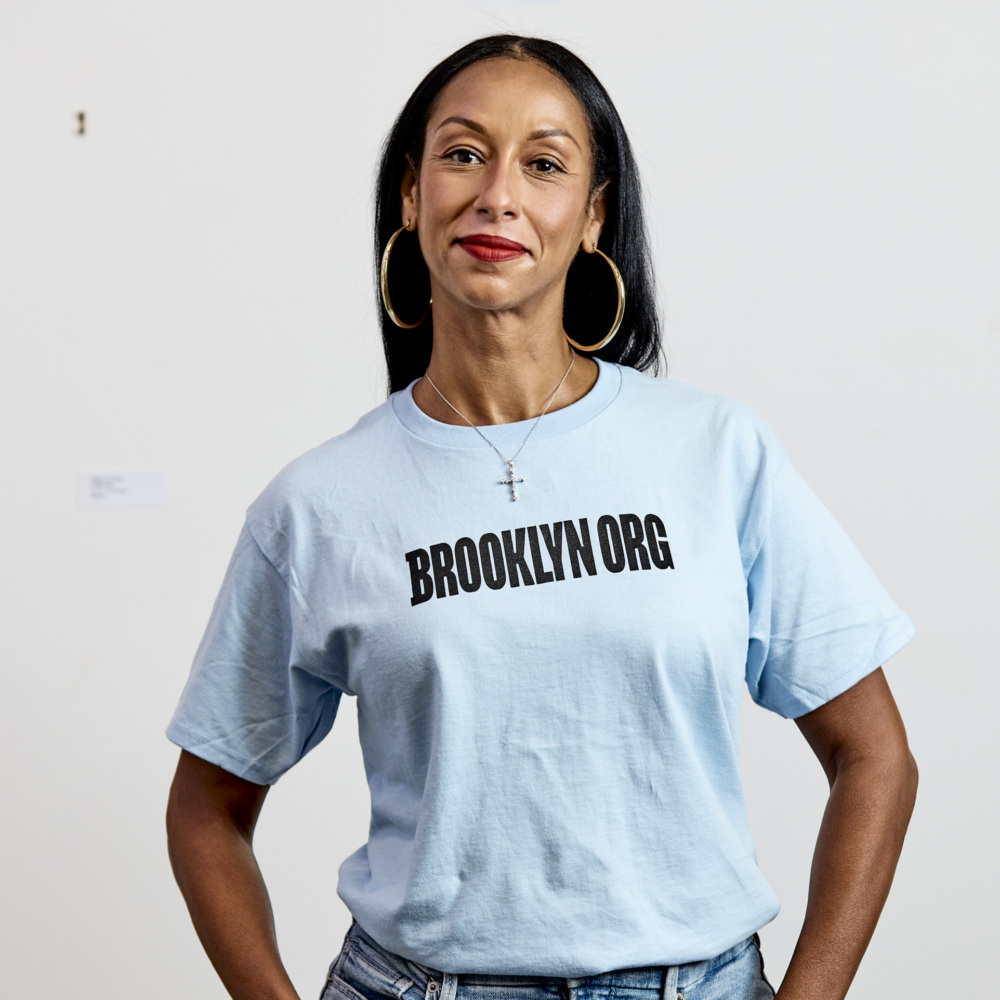 Woman wearing a light blue "BROOKLYN ORG" t-shirt, blue jeans, and hoop earrings, standing against a plain white background.