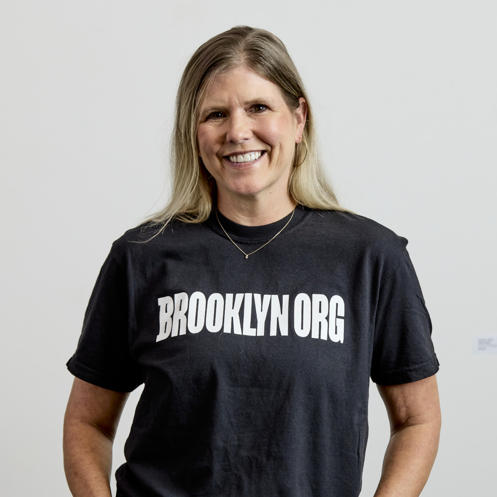 A woman with long blonde hair wearing a "BROOKLYN ORG" black t-shirt and blue jeans stands smiling against a plain white background.