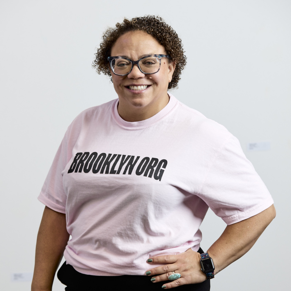 Person wearing glasses and a light pink "BROOKLYN.ORG" t-shirt, standing against a plain white background and smiling at the camera.