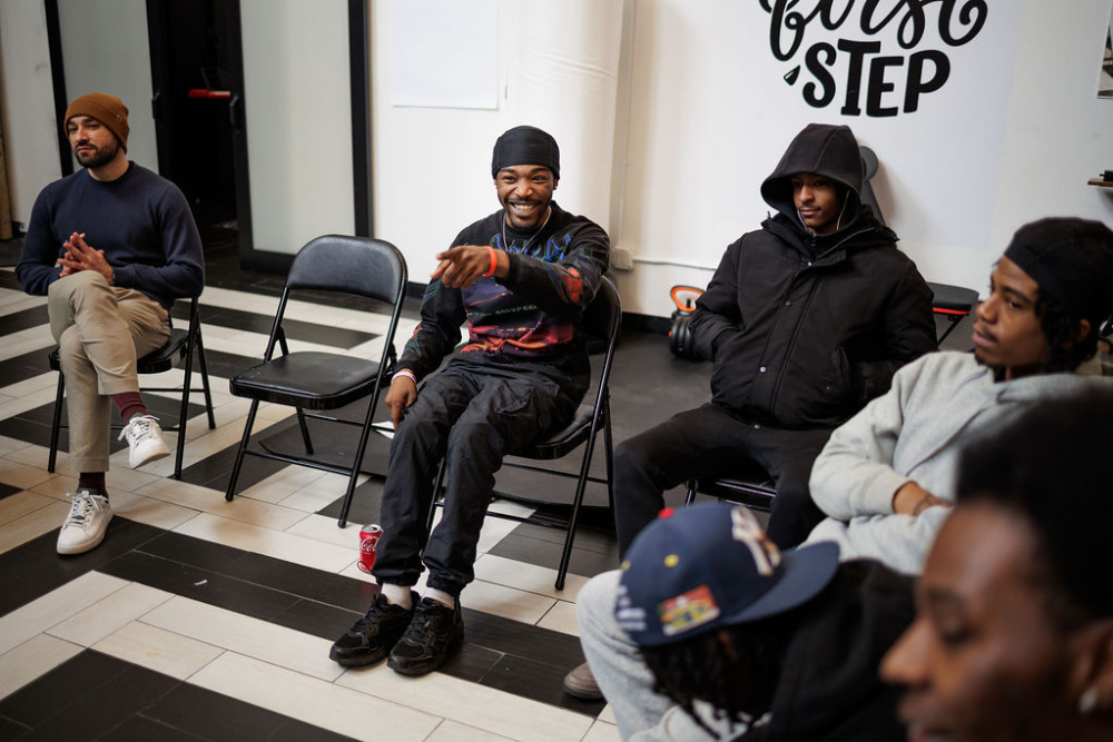 A group of men sit on folding chairs in a room, some engaged in conversation, one man smiling and pointing. The wall behind them has the words "First Step.