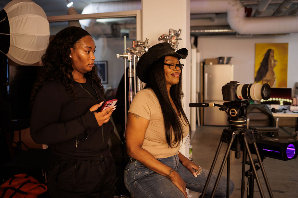 Two women in a photo studio, one standing holding a phone and the other sitting on a stool wearing a cowboy hat, next to a camera on a tripod.