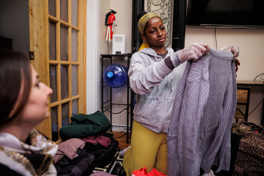A woman wearing gloves examines a gray quilted garment indoors, with folded clothes and another seated person nearby.