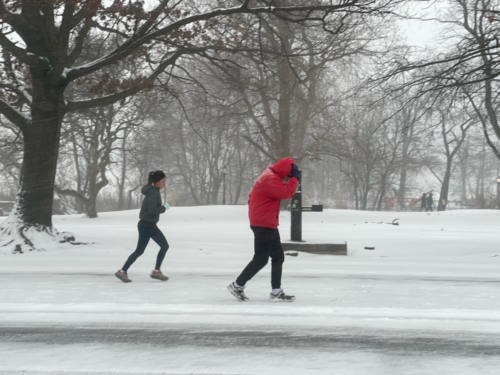 Two people jog on a snow-covered road in a park during winter, with bare trees and falling snow visible in the background.