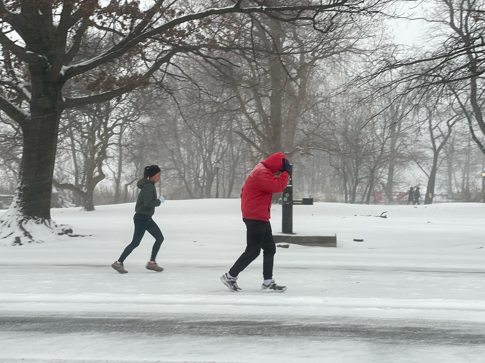 Two people jog on a snow-covered road in a park during winter, with bare trees and falling snow visible in the background.