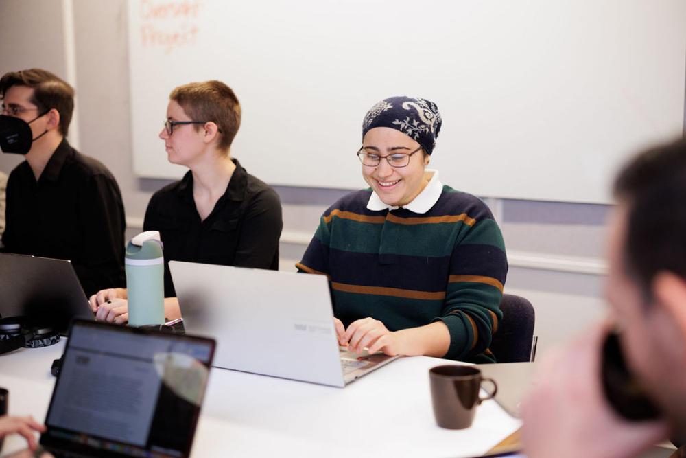 Several people sit at a table with laptops and notebooks, working and conversing in a meeting room. One person in the center is smiling while typing on their laptop.