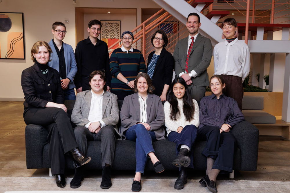 Eleven people, dressed in business casual attire, pose for a group photo indoors with some seated on a dark couch and others standing behind in front of a staircase.