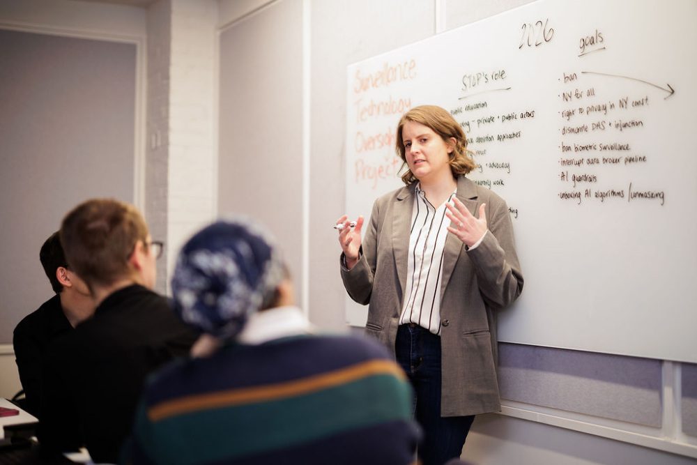 A woman stands in front of a whiteboard giving a presentation to a small group; the whiteboard has handwritten notes about surveillance technology and project goals.