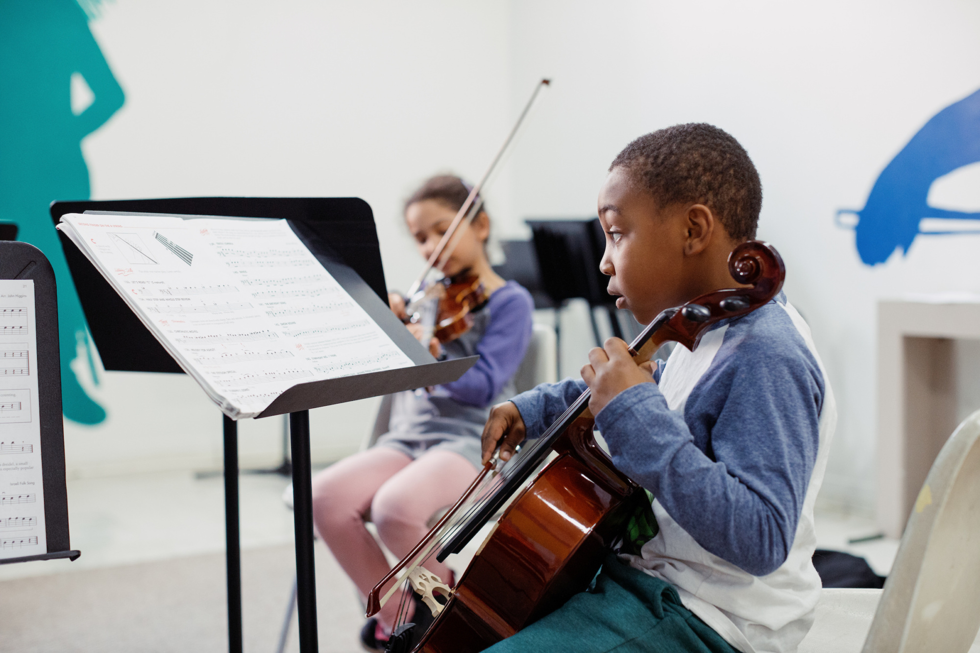 A boy plays the cello while a girl plays the violin in a music classroom, both reading sheet music from stands in front of them.
