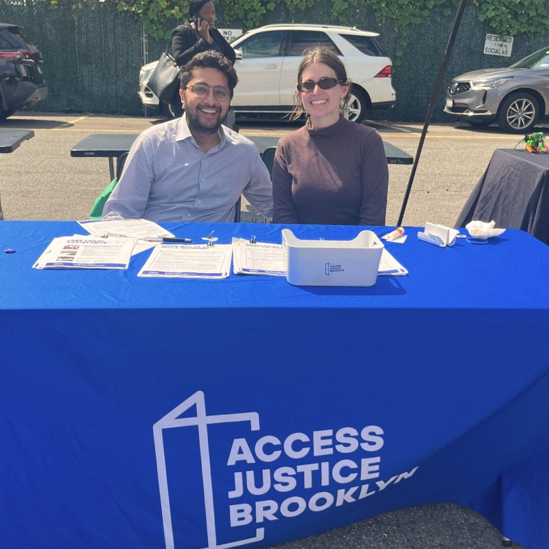 Two people sit at an outdoor table covered with a blue "Access Justice Brooklyn" tablecloth, with forms and informational materials laid out in front of them.