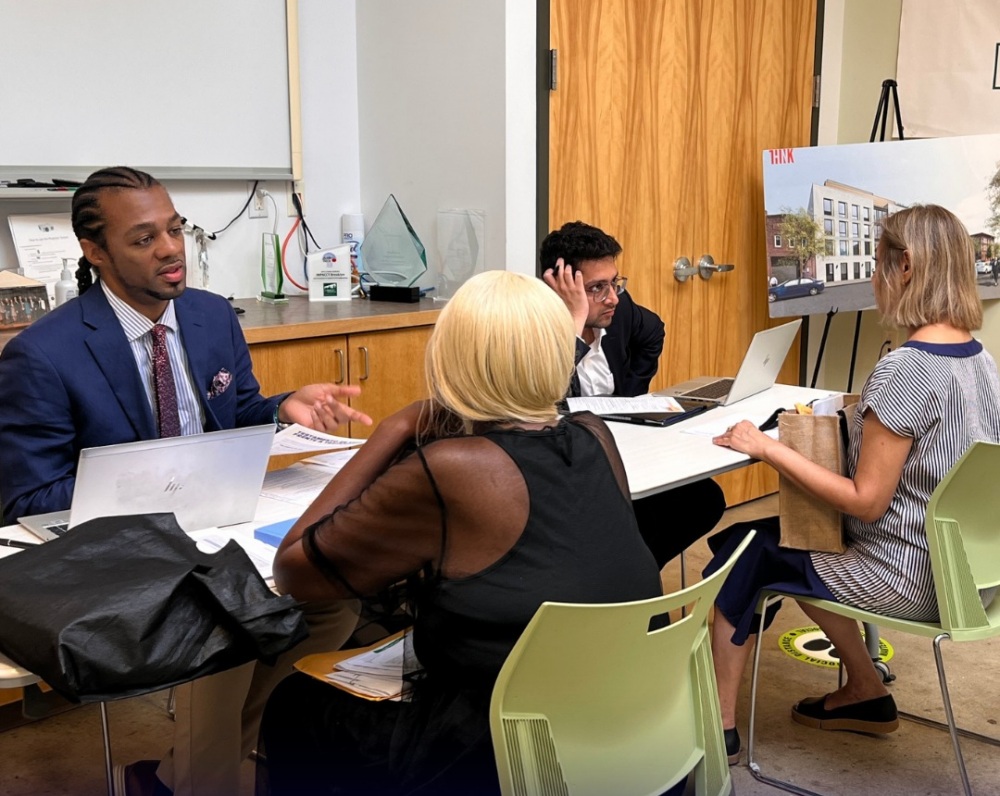 Four people in business attire sit around a table with laptops and documents, having a discussion in a modern office meeting room. A building rendering is displayed on an easel nearby.