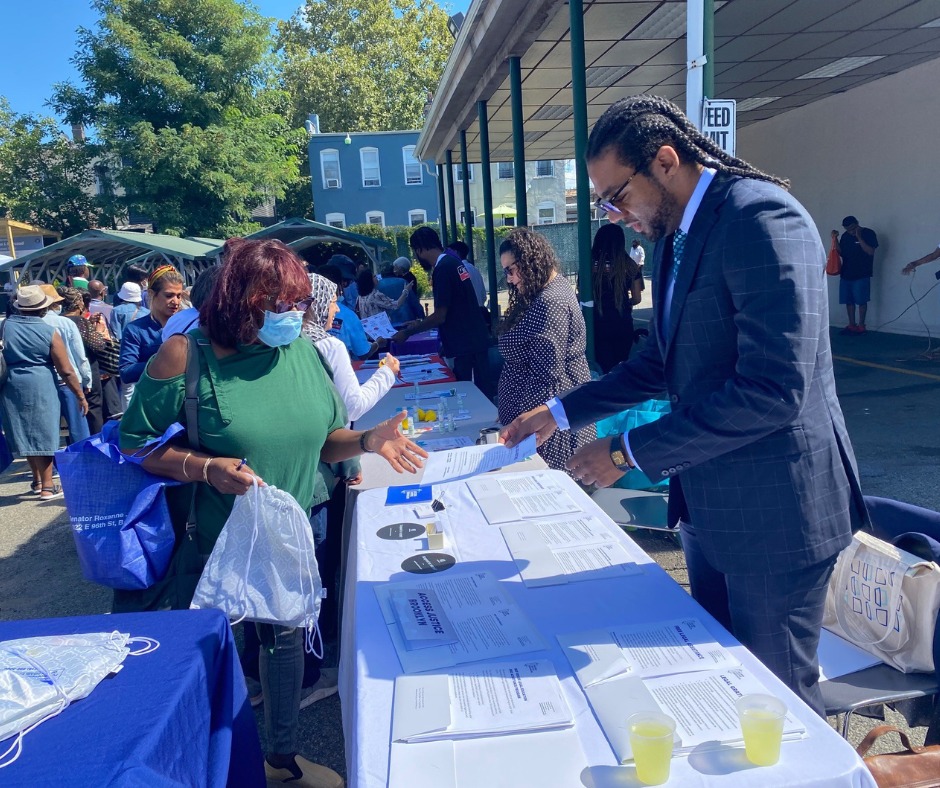 People interact at an outdoor information fair, standing at tables with documents and flyers. One man hands a paper to a woman wearing a face mask.