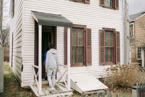 Two people in gray clothing walk into a white wooden house with brown shutters and a small porch. A ramp and leafless shrub are visible beside the entrance.