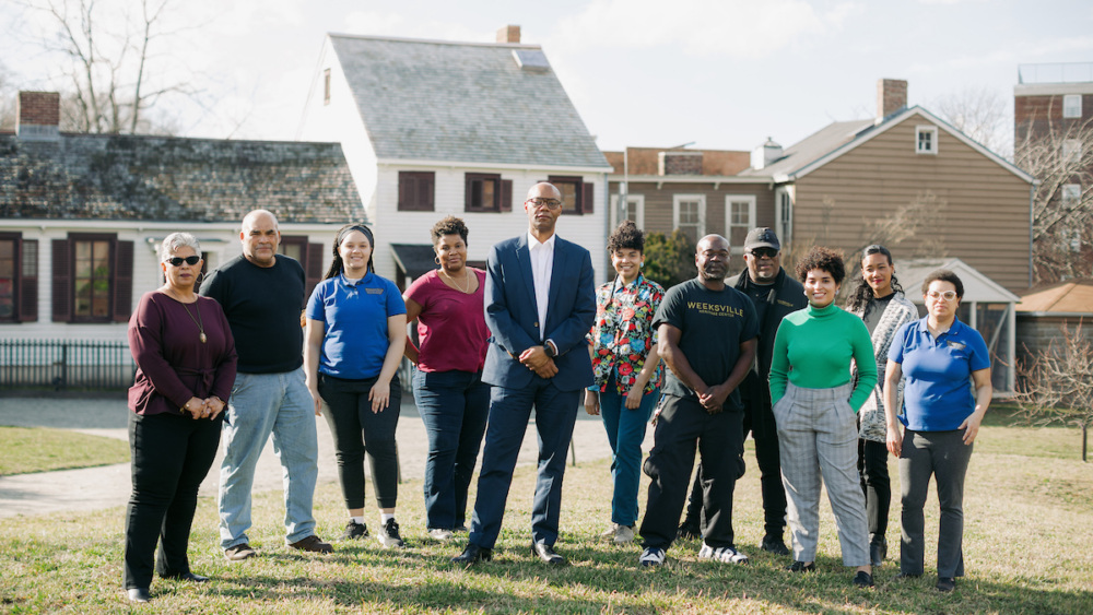 A group of eleven people stands outdoors on grass in front of historic buildings, posing for a group photo on a sunny day.