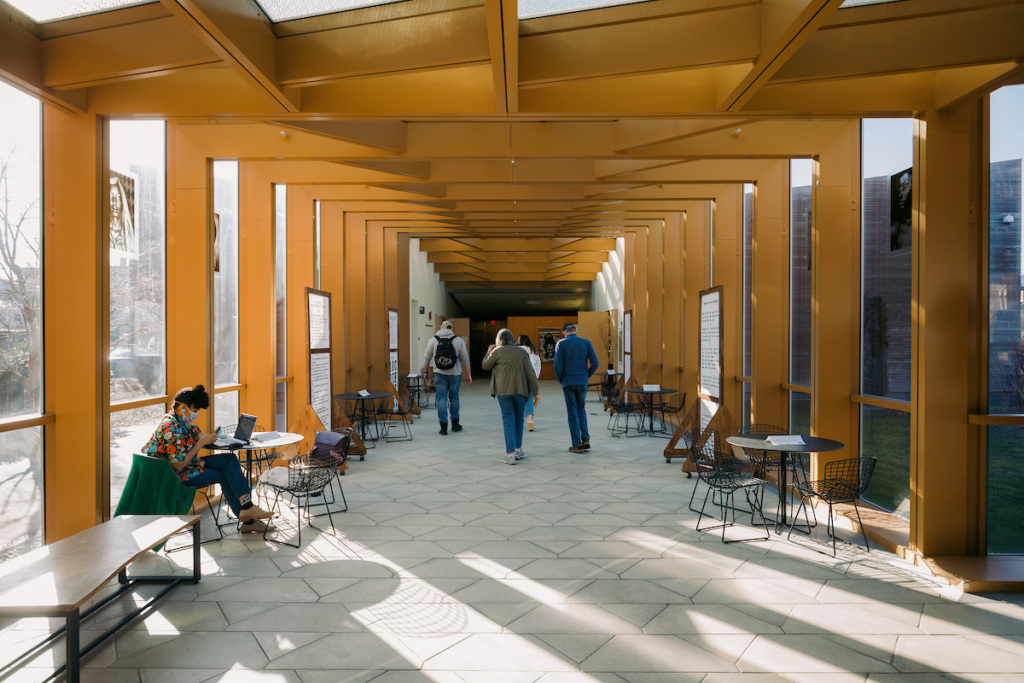 People walk down a bright, yellow corridor with glass walls and scattered tables and chairs; sunlight streams in, and a person sits alone working at a table on the left.