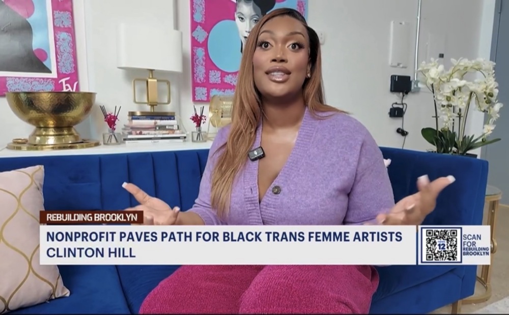 A woman in a purple sweater and pink pants sits on a blue couch, speaking, with a news headline about a nonprofit supporting Black trans femme artists displayed below.