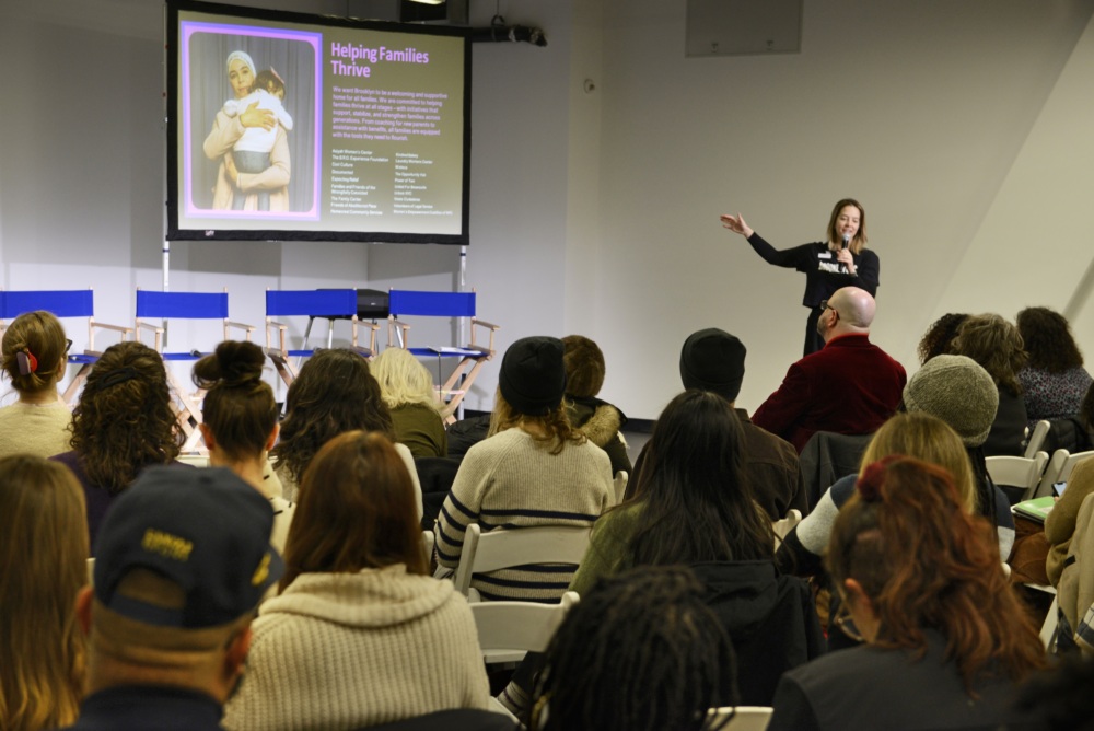 A woman gives a presentation to a seated audience, with a slide titled "Helping Families Thrive" displayed on a screen behind her.