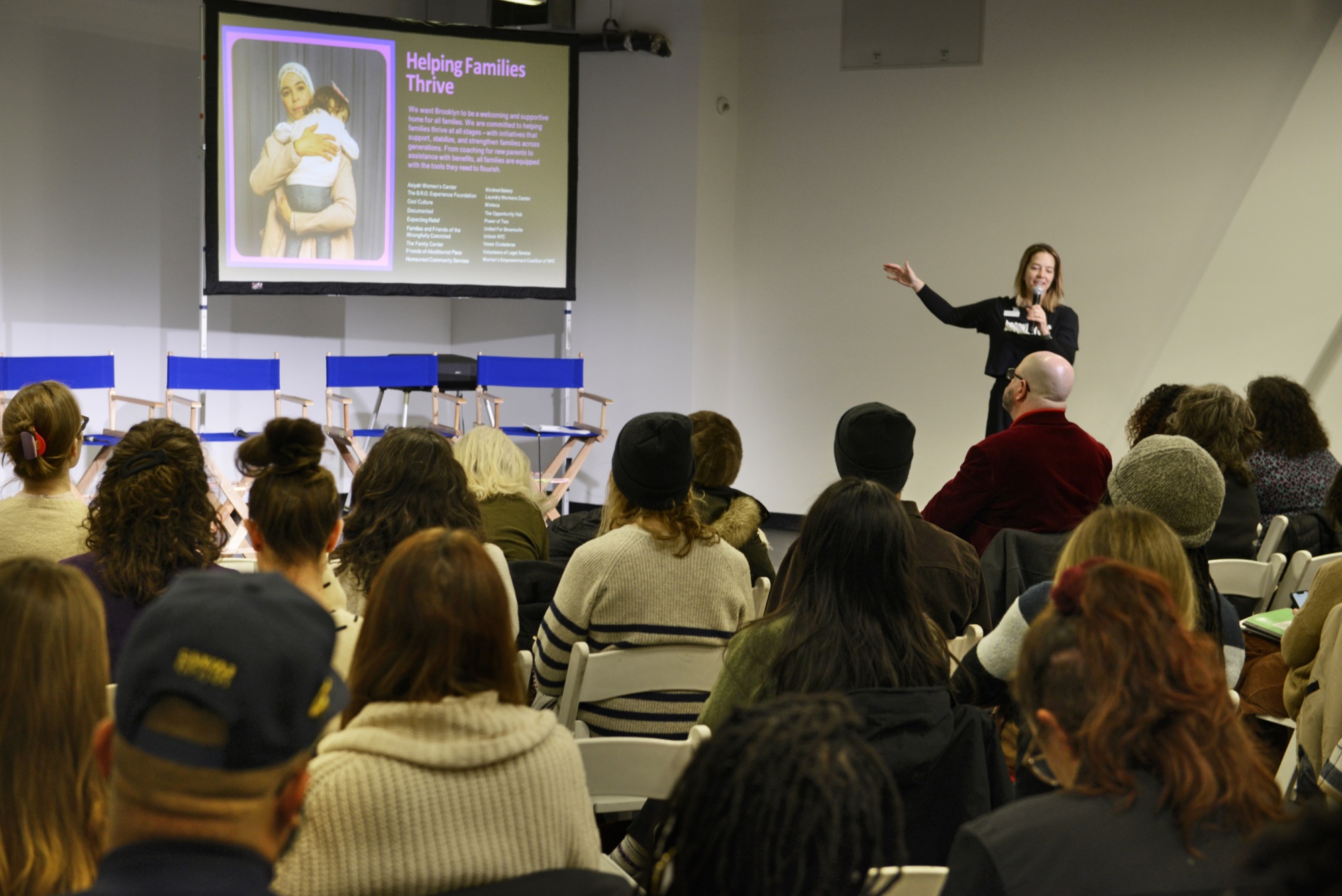 A woman gives a presentation to a seated audience, with a slide titled "Helping Families Thrive" displayed on a screen behind her.