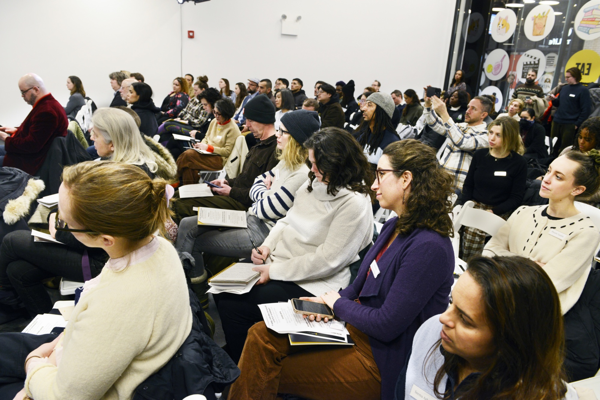 A diverse group of people sits in rows at an indoor event, listening attentively and taking notes. Some hold papers or notebooks; one person in the back is taking a photo.