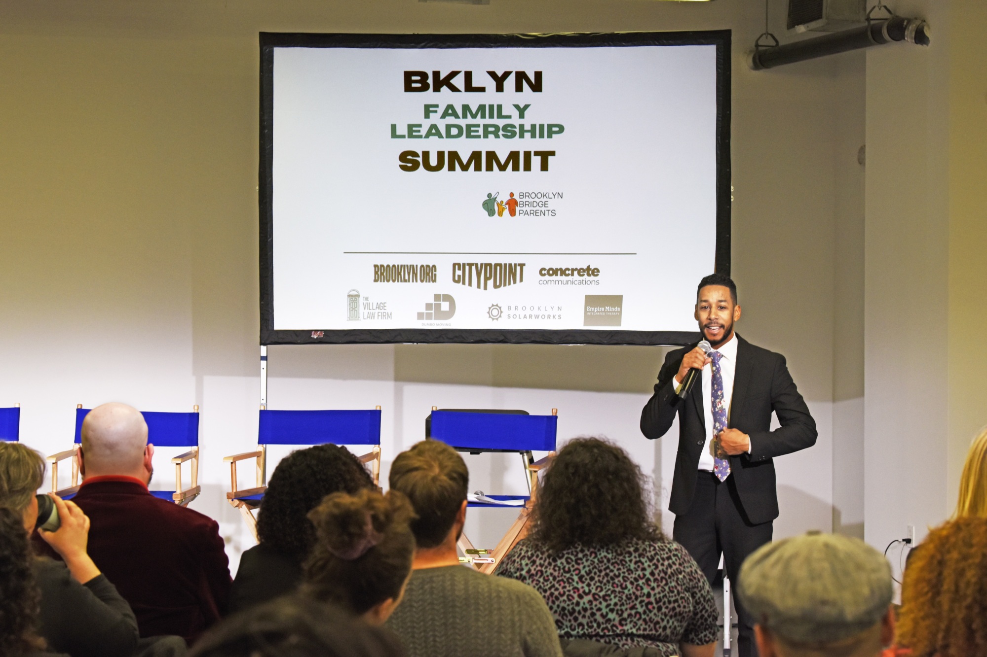 A speaker addresses an audience at the BKLYN Family Leadership Summit, with event sponsors displayed on a screen behind him.