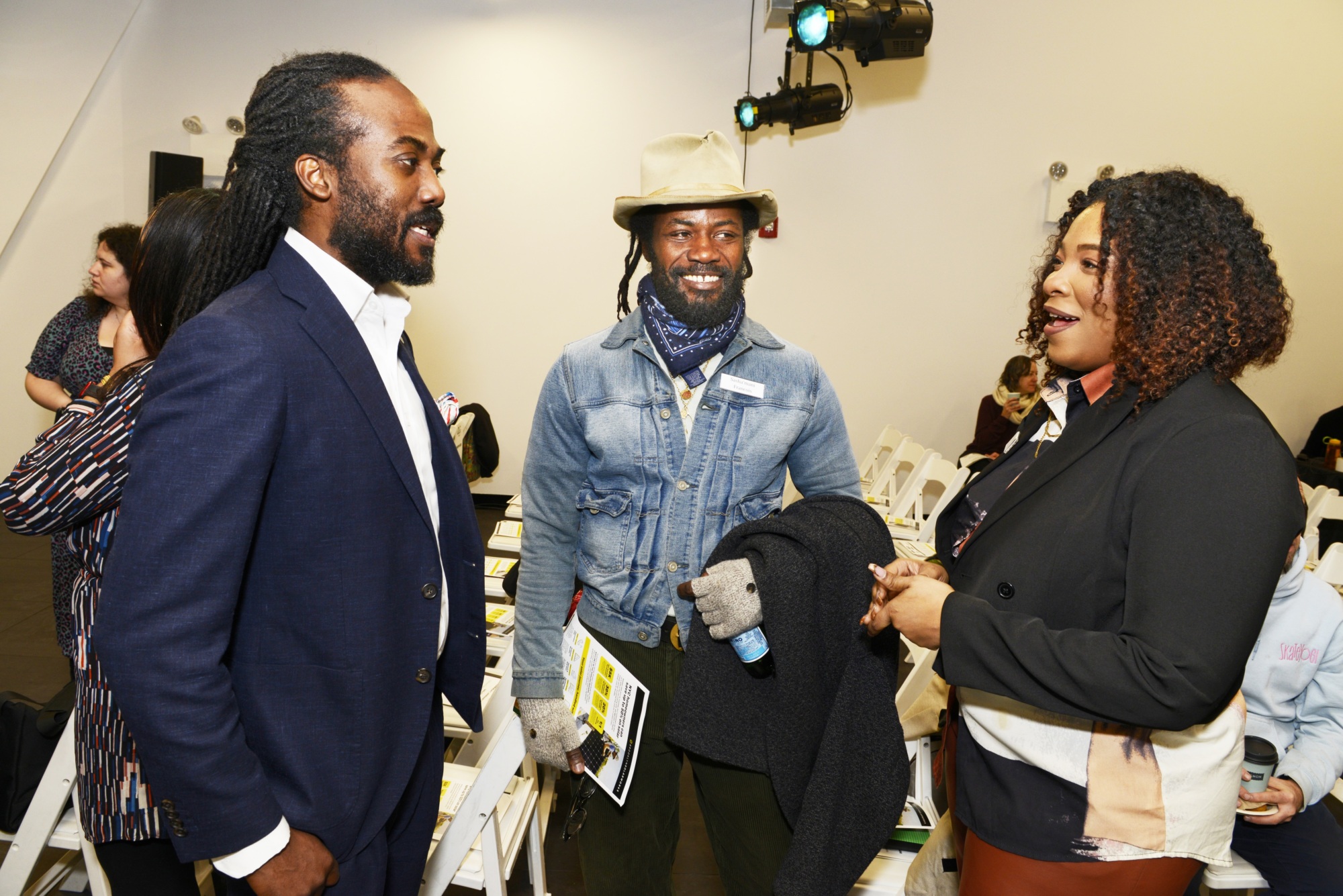 Three people stand and converse in a well-lit indoor setting, with other attendees and white folding chairs visible in the background.