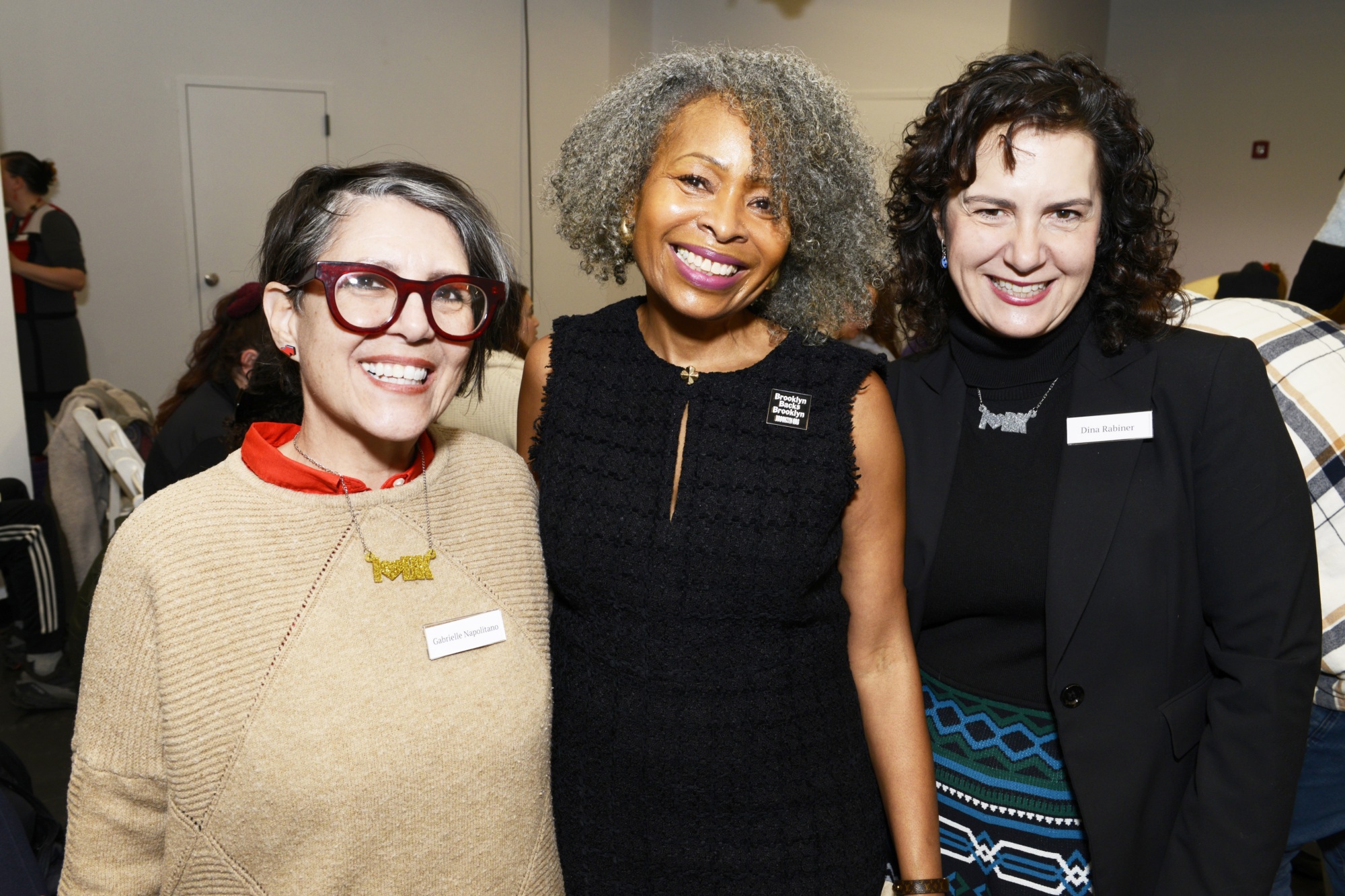 Three women stand together indoors, smiling at the camera. Two wear name tags and one wears a sleeveless black dress, while the others are in sweaters and blazers.