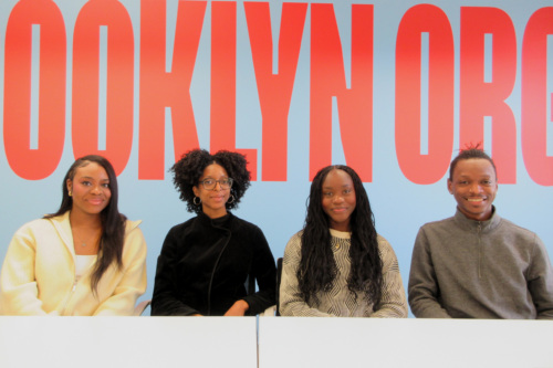 Four people sit side by side in front of a blue wall with large red text partially reading "BROOKLYN ORG.