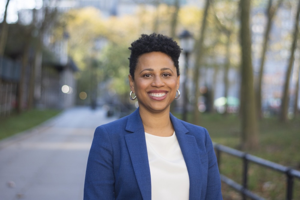 A person in a blue blazer and white shirt smiles while standing on a tree-lined city sidewalk during daytime.