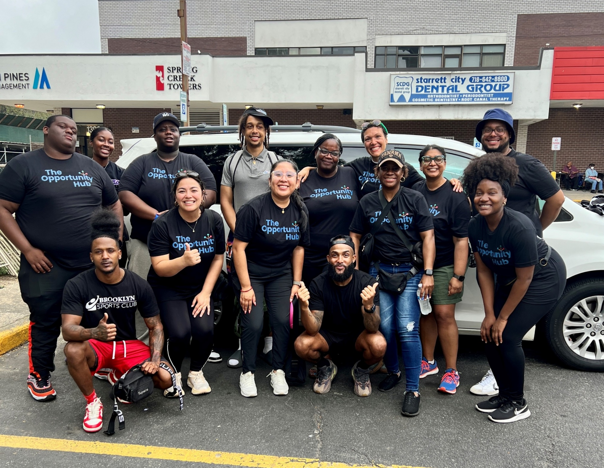 A group of people poses and smiles together outside a building, many wearing matching "The Opportunity Hub" shirts. A car is parked behind them, and storefronts are visible in the background.