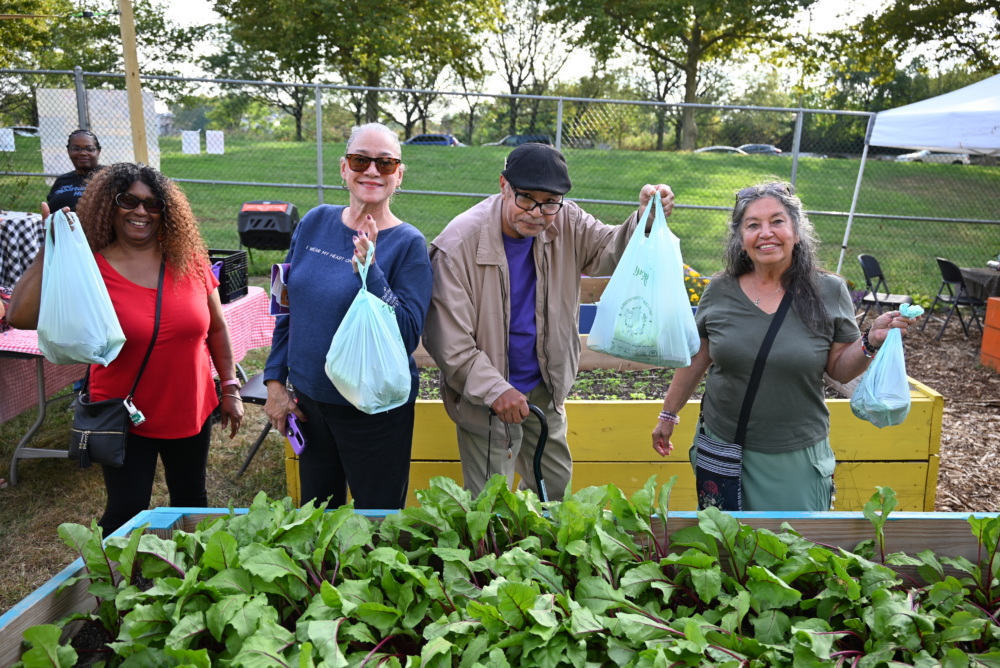 Four adults stand behind a raised garden bed holding up plastic bags, likely filled with harvested produce, at an outdoor community garden event.
