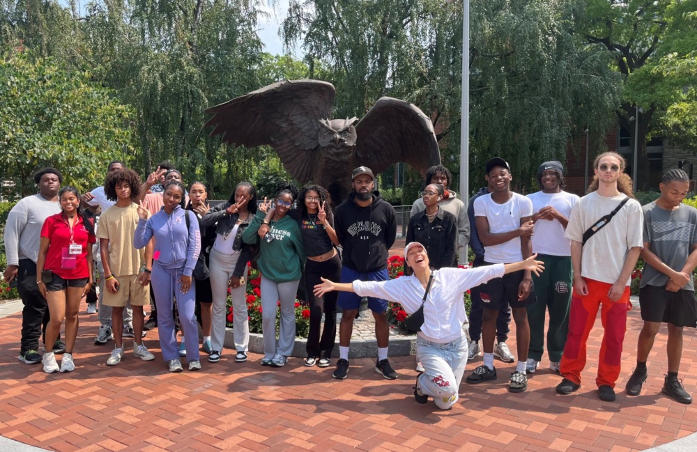 A group of people pose together in front of a large eagle statue outdoors, standing on a brick pathway surrounded by greenery and flowers.