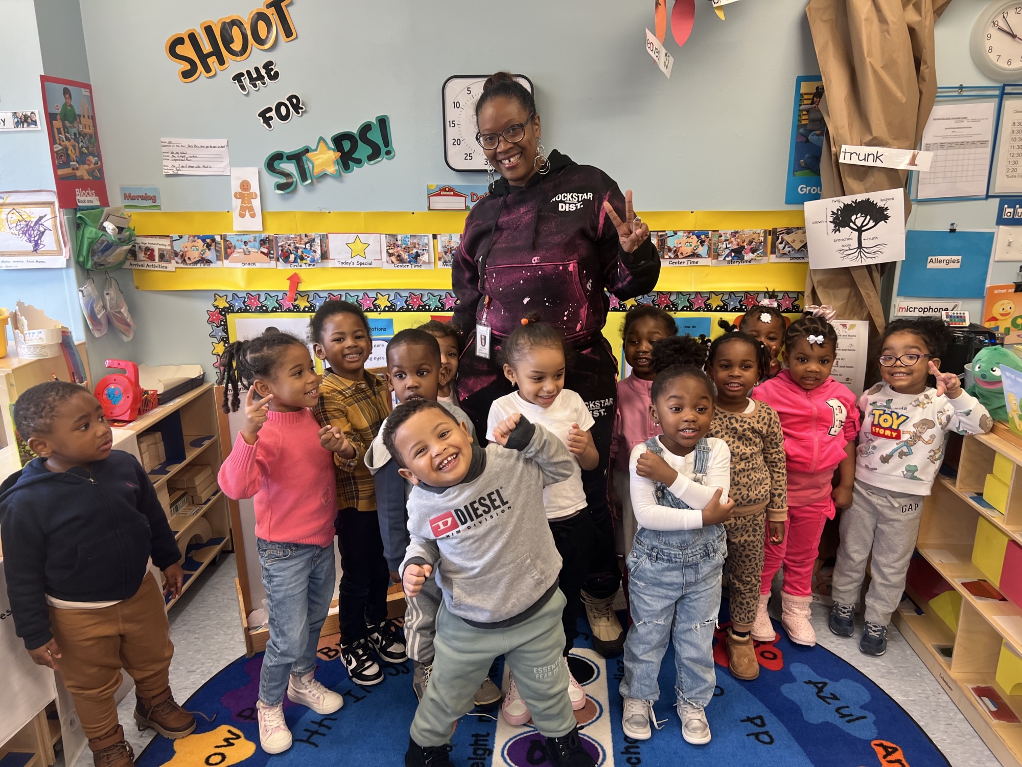 A teacher stands with a group of young children posing and smiling in a colorful classroom decorated with artwork and educational posters.