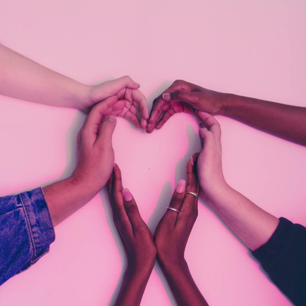Four hands of varying skin tones form the shape of a heart on a pink background.