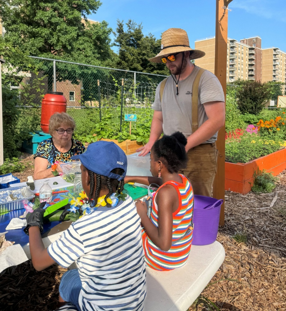 A man in a straw hat stands by a table with two children and an older woman in a community garden on a sunny day.